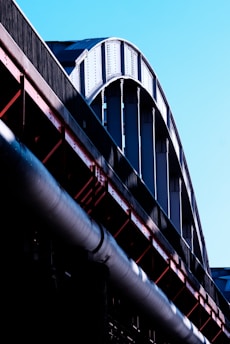 A close-up view of a large steel structure, possibly part of a bridge or industrial building, with curved beams and metal girders in shades of silver and black. The structure contrasts against a clear blue sky, and the light emphasizes the texture and rivets of the metal.