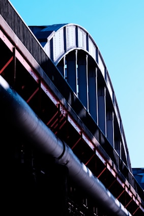 A close-up view of a large steel structure, possibly part of a bridge or industrial building, with curved beams and metal girders in shades of silver and black. The structure contrasts against a clear blue sky, and the light emphasizes the texture and rivets of the metal.