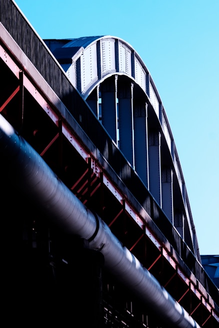 A close-up view of a large steel structure, possibly part of a bridge or industrial building, with curved beams and metal girders in shades of silver and black. The structure contrasts against a clear blue sky, and the light emphasizes the texture and rivets of the metal.