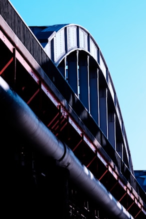 A close-up view of a large steel structure, possibly part of a bridge or industrial building, with curved beams and metal girders in shades of silver and black. The structure contrasts against a clear blue sky, and the light emphasizes the texture and rivets of the metal.