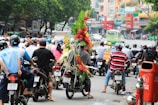 A busy urban street scene with numerous motorcyclists wearing helmets. In the center, one motorbike is adorned with large vibrant floral arrangements. The background shows a bustling city environment with various commercial signs and buildings.