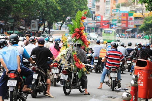 A busy urban street scene with numerous motorcyclists wearing helmets. In the center, one motorbike is adorned with large vibrant floral arrangements. The background shows a bustling city environment with various commercial signs and buildings.