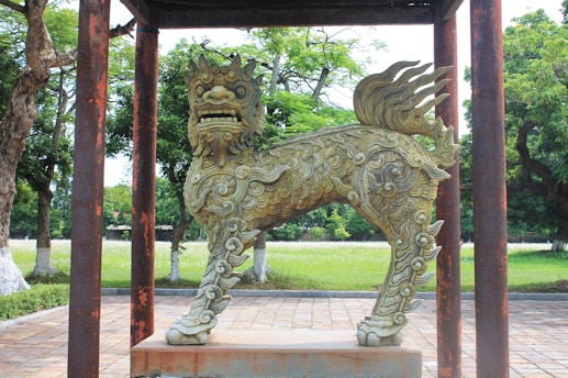 A detailed sculpture of a mythical creature with intricate patterns stands on a stone pedestal. The statue is surrounded by a lush green park, with trees in the background and a paved pathway. The creature has a lion-like face, curly mane, and a scaly body, featuring artistic stylings typical of Eastern mythology.