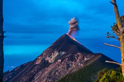 A volcano is erupting with smoke and ash billowing from its peak against a backdrop of a clear blue sky. The mountain appears rugged and dark, with patches of green vegetation visible along its slopes. Tall trees frame the scene on either side.