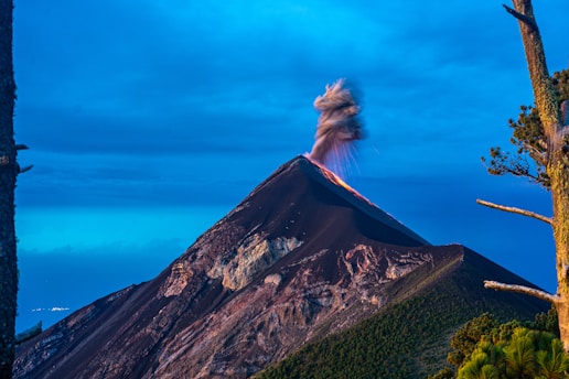 A volcano is erupting with smoke and ash billowing from its peak against a backdrop of a clear blue sky. The mountain appears rugged and dark, with patches of green vegetation visible along its slopes. Tall trees frame the scene on either side.