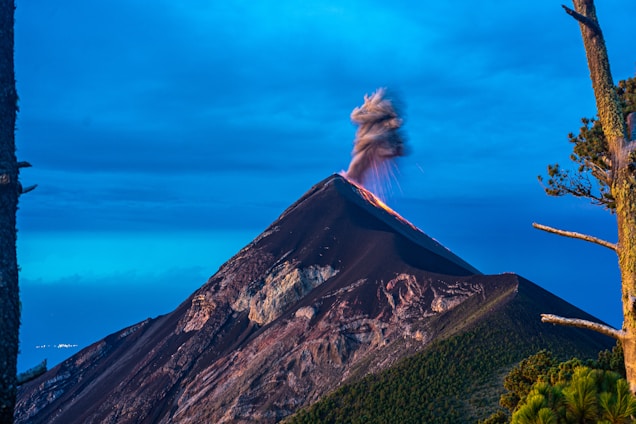 A volcano is erupting with smoke and ash billowing from its peak against a backdrop of a clear blue sky. The mountain appears rugged and dark, with patches of green vegetation visible along its slopes. Tall trees frame the scene on either side.