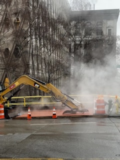An excavator is positioned on a construction site amidst a city setting. Steam or smoke rises around the machine, obscuring part of the scene. Traffic cones and barriers mark the work area, and bare trees stand in the background alongside a multi-story building.