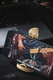 A smartphone is mounted on a snake-shaped stand, displaying a video of a person playing a bass guitar. The background features a laptop and other objects on a dark surface. The subtitle on the phone screen reads 'I CAN TELL YOU THE TRUTH'.