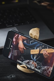 A smartphone is mounted on a snake-shaped stand, displaying a video of a person playing a bass guitar. The background features a laptop and other objects on a dark surface. The subtitle on the phone screen reads 'I CAN TELL YOU THE TRUTH'.