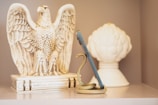 A decorative eagle sculpture is positioned to the left, with its wings spread open, crafted from a material that appears similar to ivory or ceramic. A snake-shaped phone holder is wrapped around a smartphone, positioned upright in the center. The background features a large white ornamental object, resembling a stylized artichoke or flower bud.