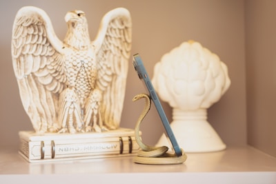 A decorative eagle sculpture is positioned to the left, with its wings spread open, crafted from a material that appears similar to ivory or ceramic. A snake-shaped phone holder is wrapped around a smartphone, positioned upright in the center. The background features a large white ornamental object, resembling a stylized artichoke or flower bud.