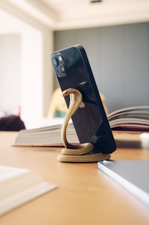 A smartphone is held upright by a snake-shaped stand on a wooden table. The stand is metallic and coiled around the base of the phone. An open book is visible in the background, with sunlight softly illuminating the scene.