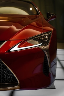A close-up view of the front section of a luxury sports car, featuring a sleek design with a shiny red finish. The intricate headlight design and the mesh grille add to the car's sophisticated appearance. The image captures the fine detailing and aerodynamic shape of the vehicle.
