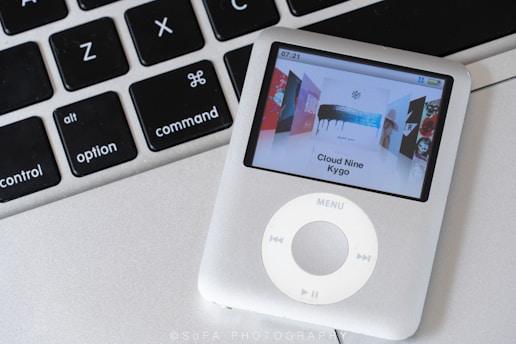 A silver iPod with a display showing the album 'Cloud Nine' by Kygo is placed on top of a silver laptop keyboard. The keyboard keys include control, option, and command keys.