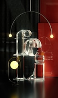 A visually striking composition featuring transparent, reflective lettering made of glass that spell out the word 'Feel.' Encircling the letters are two spherical lights, one in the foreground and another suspended from an elegant, curved arm. The background includes a textured, vertical black pattern and a contrasting red panel that add depth and contrast to the scene.