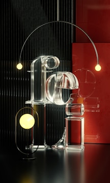 A visually striking composition featuring transparent, reflective lettering made of glass that spell out the word 'Feel.' Encircling the letters are two spherical lights, one in the foreground and another suspended from an elegant, curved arm. The background includes a textured, vertical black pattern and a contrasting red panel that add depth and contrast to the scene.