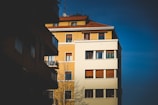 A multi-story residential building with a combination of yellow and white exterior walls and numerous windows. The building's red-tiled roof contrasts with the clear blue sky. A bare tree stands in the foreground, and shadows cover portions of the building.