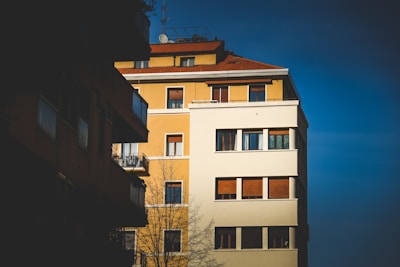 A multi-story residential building with a combination of yellow and white exterior walls and numerous windows. The building's red-tiled roof contrasts with the clear blue sky. A bare tree stands in the foreground, and shadows cover portions of the building.