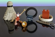 A collection of miscellaneous objects on a reflective surface, including a shuttlecock, a headless figurine with jointed arms and legs, a small flashlight, a tire-shaped object with a metal component, a red building block piece inside a bracelet with 'Love' inscribed, and a feather-shaped metallic object.