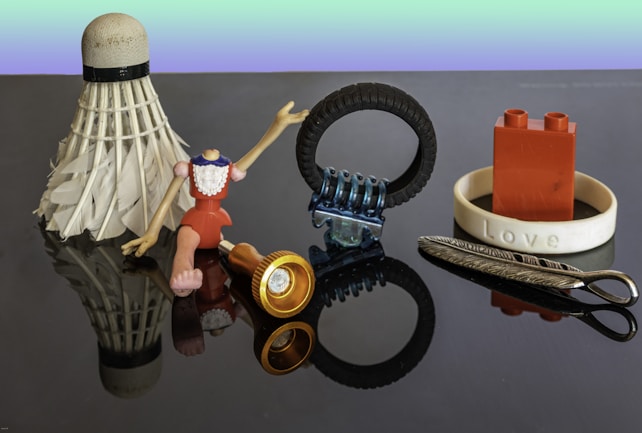 A collection of miscellaneous objects on a reflective surface, including a shuttlecock, a headless figurine with jointed arms and legs, a small flashlight, a tire-shaped object with a metal component, a red building block piece inside a bracelet with 'Love' inscribed, and a feather-shaped metallic object.
