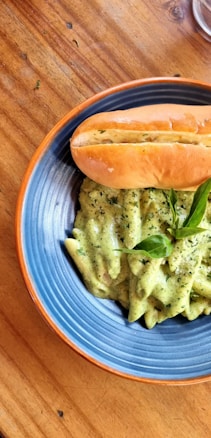 A blue bowl filled with creamy green pasta garnished with herbs and topped with a small baguette. The bowl is placed on a wooden table, and there is a sprig of fresh basil on top of the pasta.