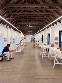 A gallery space with a high wooden ceiling and long tables displaying various documents and photographs. A person is sitting on a bench on the left, engaged with a mobile device, while another person is viewing the exhibits.