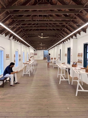 A gallery space with a high wooden ceiling and long tables displaying various documents and photographs. A person is sitting on a bench on the left, engaged with a mobile device, while another person is viewing the exhibits.