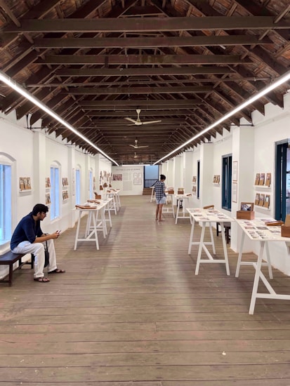 A gallery space with a high wooden ceiling and long tables displaying various documents and photographs. A person is sitting on a bench on the left, engaged with a mobile device, while another person is viewing the exhibits.
