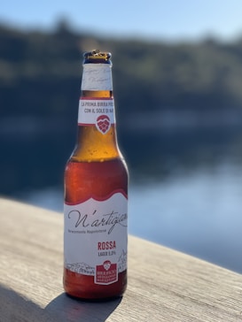 A bottle of N'artigiana beer is placed on a wooden surface outdoors with a blurred natural background. The label on the bottle indicates it is a 'Rossa' lager with 5% alcohol content, made by Birrificio Artigianale Napoletano.