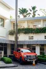 A bright orange pickup truck is parked in front of a white building with striped blue and white awnings. The setting includes lush green bushes and tall palm trees, which add a tropical feel. A traffic cone is placed in front of the truck, suggesting a reserved or restricted parking spot.