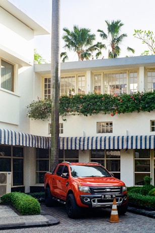 A bright orange pickup truck is parked in front of a white building with striped blue and white awnings. The setting includes lush green bushes and tall palm trees, which add a tropical feel. A traffic cone is placed in front of the truck, suggesting a reserved or restricted parking spot.