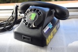 A vintage rotary dial telephone with a sticker featuring a stylized illustration of a woman's face on the dial. The phone is black and appears to be resting on a magazine or newspaper. There are additional stickers on the phone, including one that reads 'make stickers' and another with the word 'KINETIC' in yellow. The phone cord is coiled and extends to the left.
