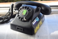 A vintage rotary dial telephone with a sticker featuring a stylized illustration of a woman's face on the dial. The phone is black and appears to be resting on a magazine or newspaper. There are additional stickers on the phone, including one that reads 'make stickers' and another with the word 'KINETIC' in yellow. The phone cord is coiled and extends to the left.