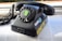 A vintage rotary dial telephone with a sticker featuring a stylized illustration of a woman's face on the dial. The phone is black and appears to be resting on a magazine or newspaper. There are additional stickers on the phone, including one that reads 'make stickers' and another with the word 'KINETIC' in yellow. The phone cord is coiled and extends to the left.