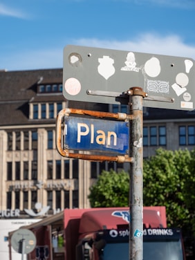 A street sign with the word 'Plan' displayed, attached to a rusty pole. Several stickers adorn the back of the sign. A large building with a flat roof is in the background, along with partially visible trees and a red truck parked nearby.