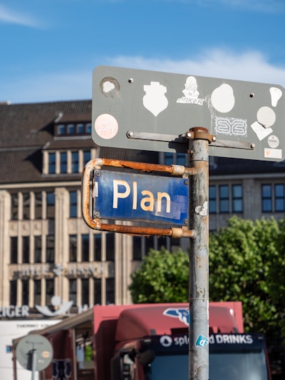 A street sign with the word 'Plan' displayed, attached to a rusty pole. Several stickers adorn the back of the sign. A large building with a flat roof is in the background, along with partially visible trees and a red truck parked nearby.