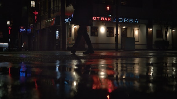 A dimly lit urban scene showing a wet street at night. A person is walking past a neon-lit bar called Zorba, with reflections of the signage and lights visible on the wet pavement. Additional neon signs and a few lit windows add to the ambiance of the empty street.