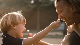 A child and an adult are playfully interacting, with the child holding a small flower up to the adult's nose. Both are smiling warmly, and there is a feeling of affection between them. The background appears softly blurred, suggesting a natural or park-like setting.