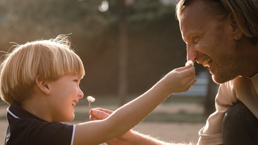 A child and an adult are playfully interacting, with the child holding a small flower up to the adult's nose. Both are smiling warmly, and there is a feeling of affection between them. The background appears softly blurred, suggesting a natural or park-like setting.