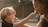 A child and an adult are playfully interacting, with the child holding a small flower up to the adult's nose. Both are smiling warmly, and there is a feeling of affection between them. The background appears softly blurred, suggesting a natural or park-like setting.