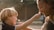 A child and an adult are playfully interacting, with the child holding a small flower up to the adult's nose. Both are smiling warmly, and there is a feeling of affection between them. The background appears softly blurred, suggesting a natural or park-like setting.