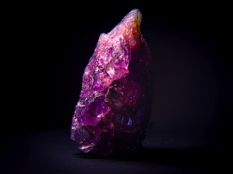 A large, rough amethyst crystal with a vibrant purple hue is displayed against a black background. The crystal features complex facets that catch the light, revealing shades of purple, violet, and slight pinkish tones. Its irregular shape adds a raw, natural aesthetic.