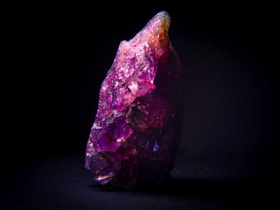 A large, rough amethyst crystal with a vibrant purple hue is displayed against a black background. The crystal features complex facets that catch the light, revealing shades of purple, violet, and slight pinkish tones. Its irregular shape adds a raw, natural aesthetic.