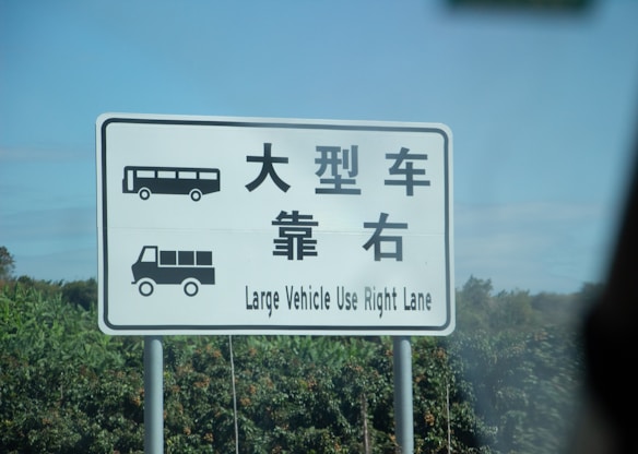 A road sign with both English and Chinese characters instructs large vehicles to use the right lane. It includes symbols of a bus and a truck, indicating vehicles that should follow the direction. The background consists of blurred greenery and a clear blue sky.
