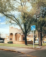 A park area features a stone structure with arches, surrounded by lush green trees and a sidewalk. A blue handicapped parking sign is visible near the foreground. Several cars are parked in the background, and a streetlamp stands nearby, adding to the urban setting.