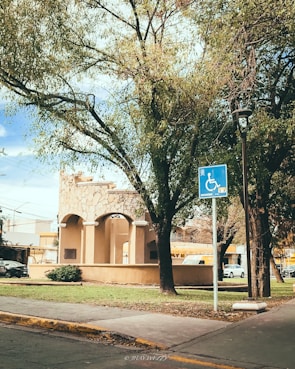 A park area features a stone structure with arches, surrounded by lush green trees and a sidewalk. A blue handicapped parking sign is visible near the foreground. Several cars are parked in the background, and a streetlamp stands nearby, adding to the urban setting.