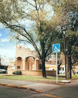 A park area features a stone structure with arches, surrounded by lush green trees and a sidewalk. A blue handicapped parking sign is visible near the foreground. Several cars are parked in the background, and a streetlamp stands nearby, adding to the urban setting.