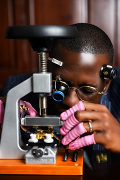 A person wearing magnifying glasses and pink gloves is carefully working on a mechanical device, possibly a watch or small machinery, using a specialized tool. The person is fully focused on the task, with a background that appears to be wooden furniture.