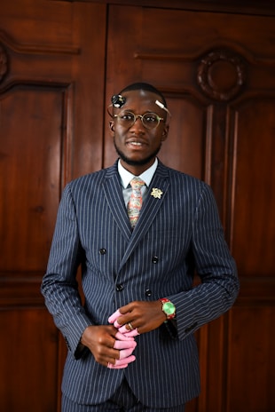 A man in a pinstripe suit and floral tie stands in front of a wooden paneled background. He is wearing glasses and has a decorative pin on his lapel. He holds a pair of folded pink gloves in his hands and has a green wristwatch. His expression is poised and serious.