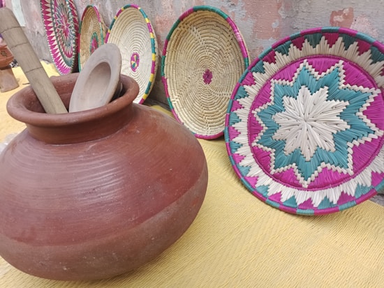 A collection of woven baskets with intricate patterns is displayed against a textured wall. The baskets feature vibrant colors like pink, teal, and natural beige. In the foreground, there is a large clay pot with wooden utensils inside, including a pestle and a flat round object.